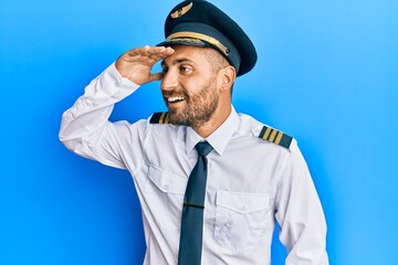 Handsome man with beard wearing airplane pilot uniform very happy and smiling looking far away with hand over head. searching concept.