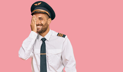Handsome man with beard wearing airplane pilot uniform covering one eye with hand, confident smile on face and surprise emotion.