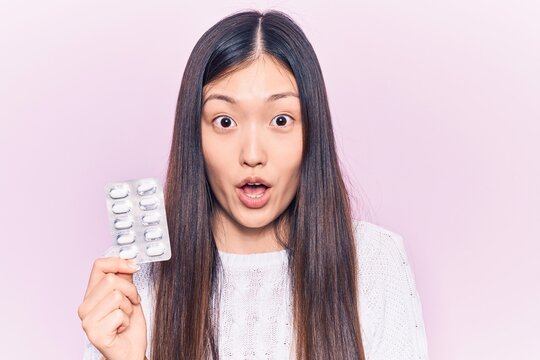 Young beautiful chinese woman holding pills scared and amazed with open mouth for surprise, disbelief face