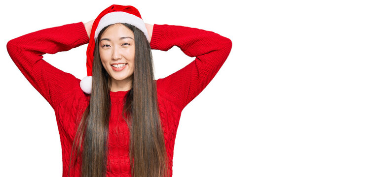 Young chinese woman wearing christmas hat relaxing and stretching, arms and hands behind head and neck smiling happy