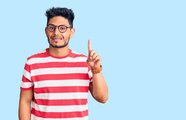 Fototapeta premium Handsome latin american young man wearing casual clothes and glasses showing and pointing up with finger number one while smiling confident and happy.
