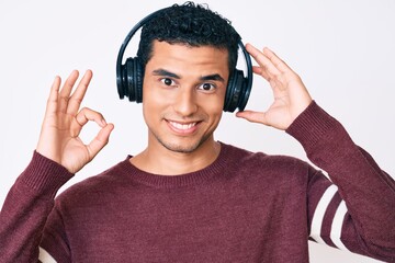 Young handsome hispanic man listening to music using headphones doing ok sign with fingers, smiling friendly gesturing excellent symbol