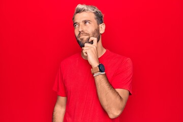 Young handsome blond man wearing casual red t-shirt standing over isolated red background thinking concentrated about doubt with finger on chin and looking up wondering
