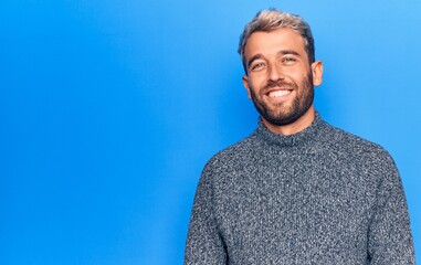 Young handsome blond man wearing casual sweater standing over isolated blue background with a happy and cool smile on face. Lucky person.