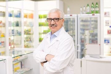 Naklejka na ściany i meble Aged drugstore worker in whitecoat and eyeglasses standing against displays