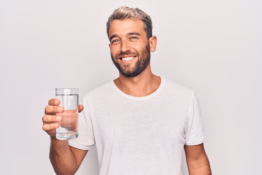 Handsome blond man with beard drinking glass of water to refreshment over white background looking positive and happy standing and smiling with a confident smile showing teeth