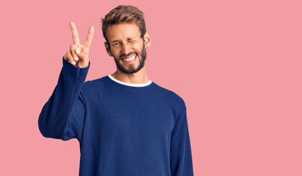 Handsome blond man with beard wearing casual sweater smiling with happy face winking at the camera doing victory sign with fingers. number two.