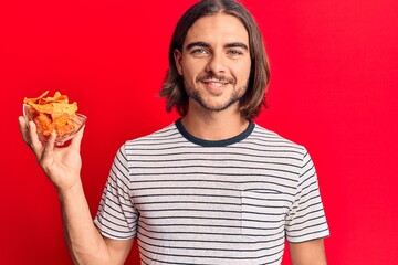 Young handsome man holding nachos potato chips looking positive and happy standing and smiling with...