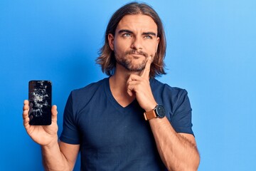 Young handsome man holding broken smartphone showing cracked screen serious face thinking about question with hand on chin, thoughtful about confusing idea