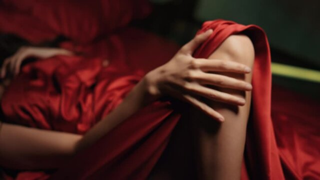 Unknown lady stroking leg in bed. Naked woman resting in red silk bed.