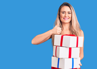 Young beautiful blonde woman holding birthday gifts smiling happy and positive, thumb up doing excellent and approval sign