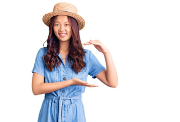 Young beautiful chinese girl wearing summer hat gesturing with hands showing big and large size sign, measure symbol. smiling looking at the camera. measuring concept.