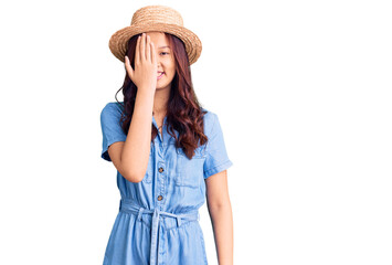 Young beautiful chinese girl wearing summer hat covering one eye with hand, confident smile on face and surprise emotion.