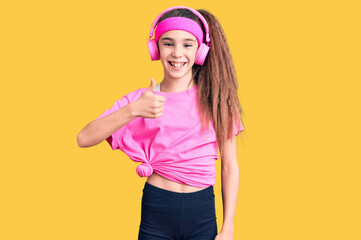 Cute hispanic child girl wearing gym clothes and using headphones doing happy thumbs up gesture with hand. approving expression looking at the camera showing success.