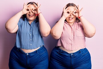 Young plus size twins wearing casual clothes doing ok gesture like binoculars sticking tongue out, eyes looking through fingers. crazy expression.