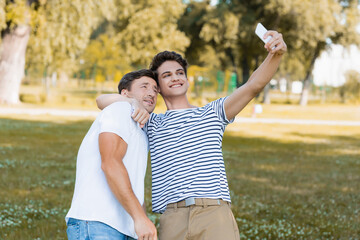 teenager son hugging father while taking selfie in green park