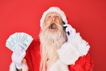 Old senior man wearing santa claus costume holding dollars and smartphone angry and mad screaming frustrated and furious, shouting with anger looking up.