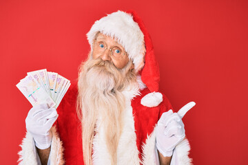 Old senior man with grey hair and long beard wearing santa claus costume holding colombian pesos smiling happy pointing with hand and finger to the side