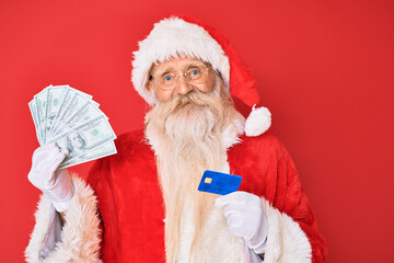 Old senior man wearing santa claus costume holding dollars and credit card smiling with a happy and cool smile on face. showing teeth.