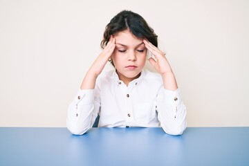 Cute hispanic child wearing casual clothes sitting on the table with hand on head, headache because stress. suffering migraine.