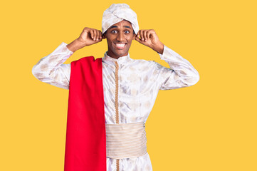 African handsome man wearing tradition sherwani saree clothes smiling pulling ears with fingers,...