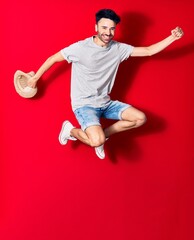 Young handsome hispanic man wearing casual clothes smiling happy. Jumping with smile on face holding summer hat celebrating with fist up over isolated red background.