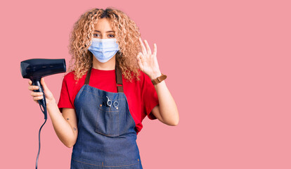 Young blonde woman with curly hair holding dryer blow wearing safety mask for coranvirus doing ok sign with fingers, smiling friendly gesturing excellent symbol