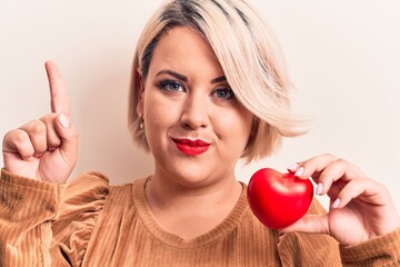 Young beautiful blonde plus size woman holding plastic heart over isolated white background smiling with an idea or question pointing finger with happy face, number one