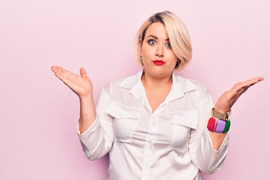 Young Beautiful Blonde Plus Size Woman Wearing Elegant Shirt Over Isolated Pink Background Clueless And Confused With Open Arms, No Idea And Doubtful Face.