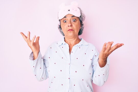 Senior hispanic grey- haired woman wearing sleep mask and pajama clueless and confused with open arms, no idea and doubtful face.