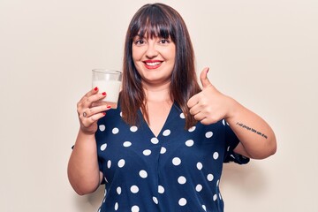 Young plus size woman holding glass of milk smiling happy and positive, thumb up doing excellent and approval sign