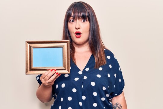 Young plus size woman holding empty frame scared and amazed with open mouth for surprise, disbelief face