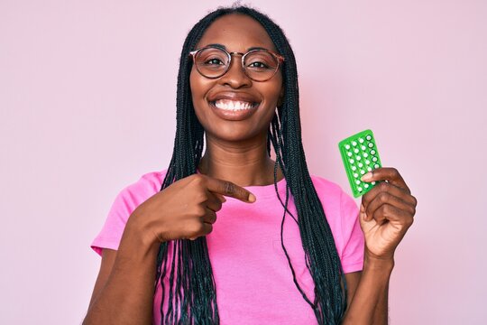 African american woman with braids holding birth control pills pointing finger to one self smiling happy and proud