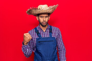 Young latin man wearing farmer hat and apron angry and mad raising fist frustrated and furious while shouting with anger. rage and aggressive concept.