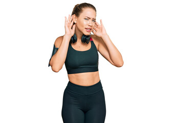 Beautiful blonde woman wearing gym clothes and using headphones trying to hear both hands on ear gesture, curious for gossip. hearing problem, deaf