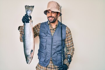 Handsome hispanic man with beard holding fishing rod and raw salmon winking looking at the camera...