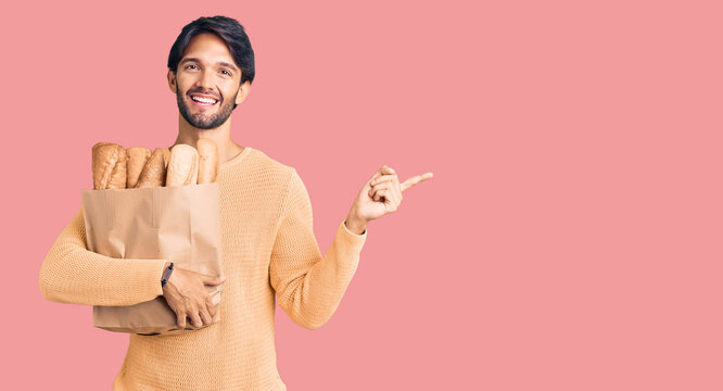 Handsome hispanic man holding paper bag with bread smiling happy pointing with hand and finger to the side