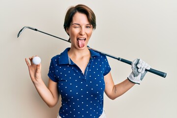 Young brunette woman with short hair holding ball and golf club sticking tongue out happy with...
