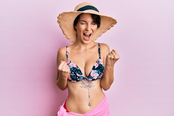 Young brunette woman with short hair wearing bikini and hat celebrating surprised and amazed for success with arms raised and eyes closed