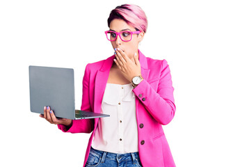 Young beautiful woman with pink hair wearing glasses holding laptop covering mouth with hand, shocked and afraid for mistake. surprised expression