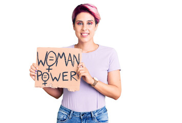 Young beautiful woman with pink hair holding woman power banner looking positive and happy standing and smiling with a confident smile showing teeth