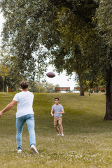 back view of father throwing rugby ball to teenager son in green park