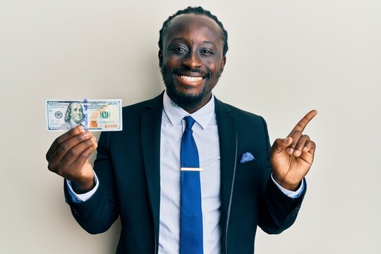 Handsome young black man wearing business suit and tie holding 100 dollars smiling happy pointing with hand and finger to the side