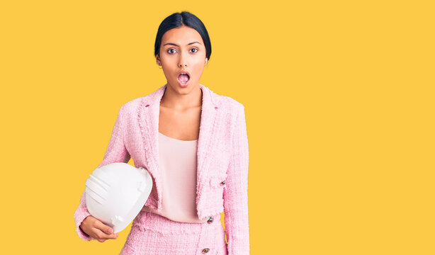 Young beautiful latin girl holding architect hardhat scared and amazed with open mouth for surprise, disbelief face