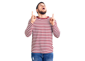 Young handsome man wearing striped sweater amazed and surprised looking up and pointing with fingers and raised arms.