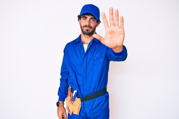 Handsome young man with curly hair and bear weaing handyman uniform doing stop sing with palm of the hand. warning expression with negative and serious gesture on the face.