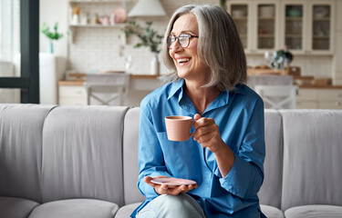 Happy beautiful relaxed mature older adult grey-haired woman drinking coffee relaxing on sofa at home. Smiling stylish middle aged 60s lady enjoying resting sitting on couch in modern living room. - Powered by Adobe