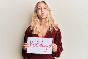 Fototapeta na wymiar Young blonde girl holding holidays banner message puffing cheeks with funny face. mouth inflated with air, catching air.