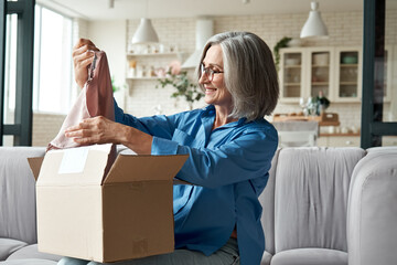 Happy middle aged woman buyer opening parcel box at home. Smiling old mature female customer...