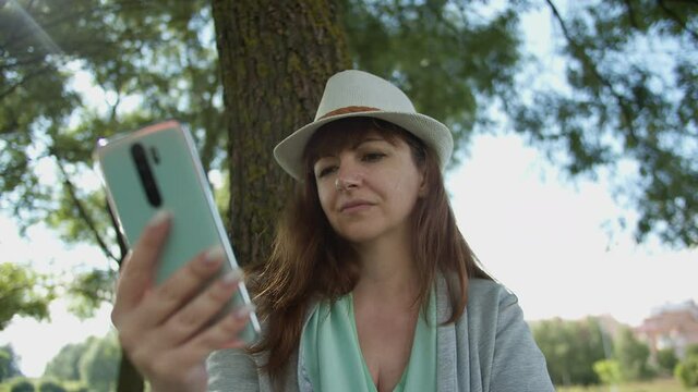 woman in a hat sits under a tree, reads some information on the phone